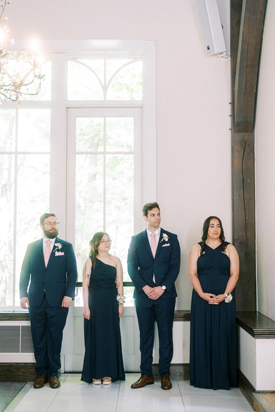Four wedding attendants in navy attire stand inside a bright room with tall windows and wood beams.