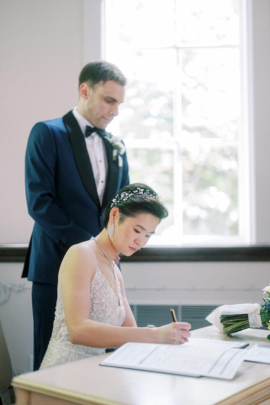 Bride signs a wedding document at a desk while the groom stands behind her in a navy tuxedo.
