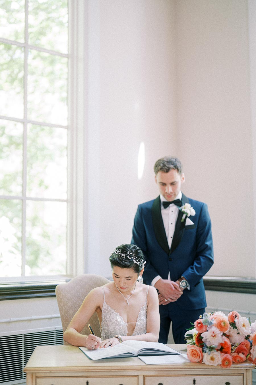 Bride signs a register at a table while groom stands behind her beside a bouquet in a bright room.