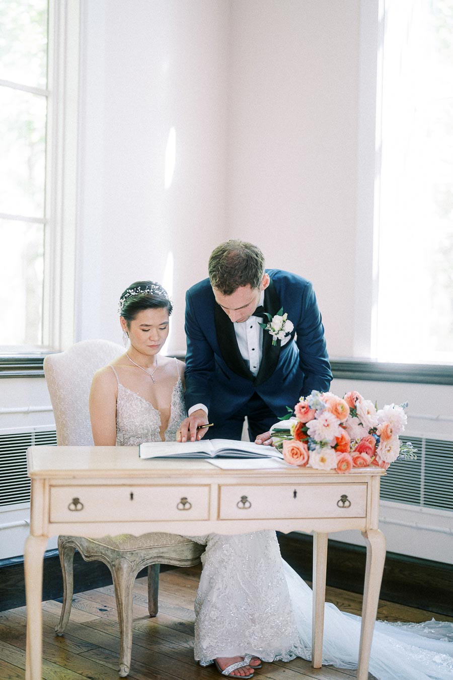 Miffy sits in her wedding gown as Rawle leans over to sign a register beside a peach floral bouquet.