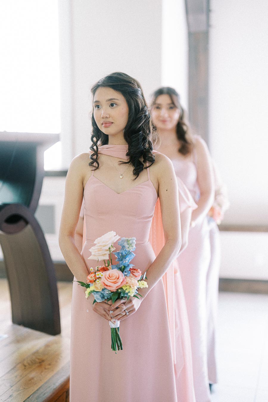 Bridesmaids in blush pink dresses holding colorful bouquets inside a bright wedding venue room.