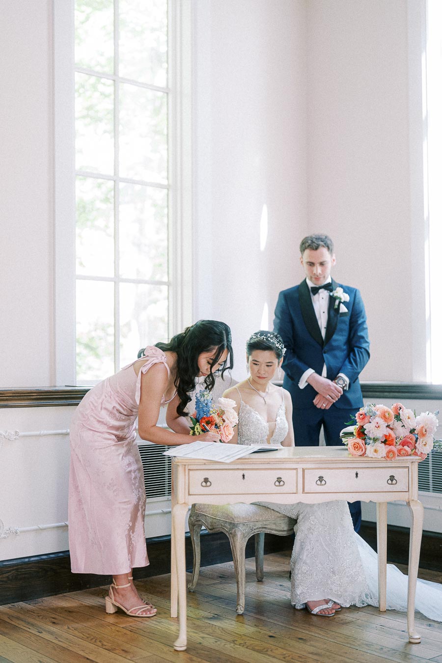 Bride signs a wedding document at a small desk while the groom and a bridesmaid stand beside her.