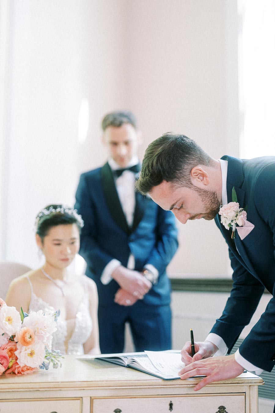 Groom signs a wedding register beside the seated bride and officiant during an indoor ceremony.