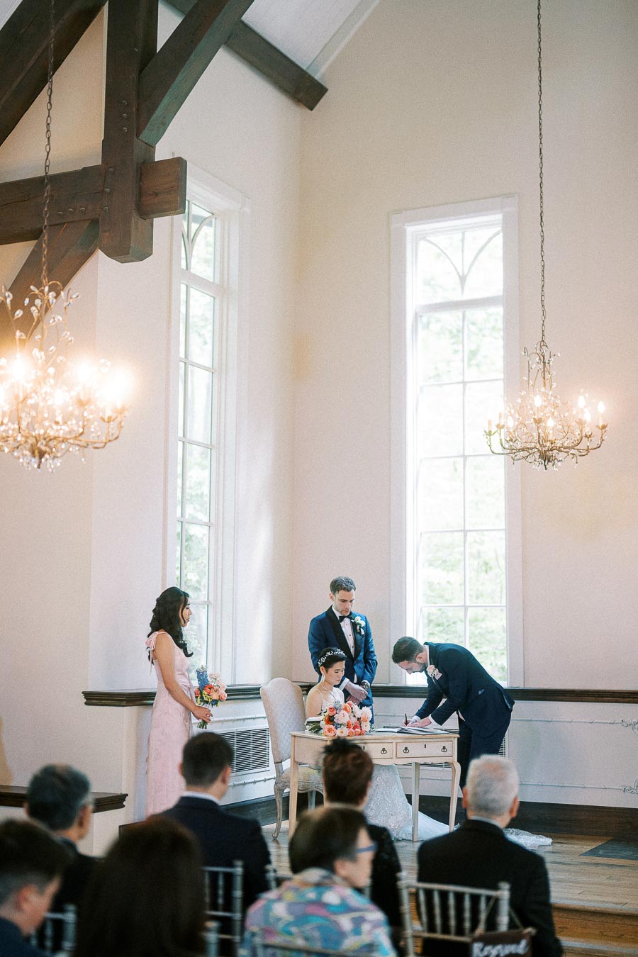 Groom signs the wedding register beside the seated bride during a bright chapel ceremony with tall windows.