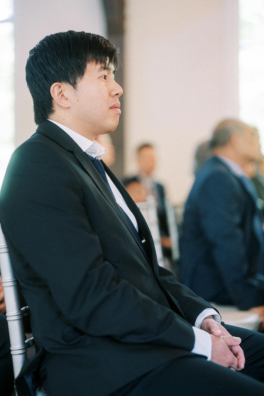 Man in a black suit sits attentively among seated guests during an indoor wedding ceremony.