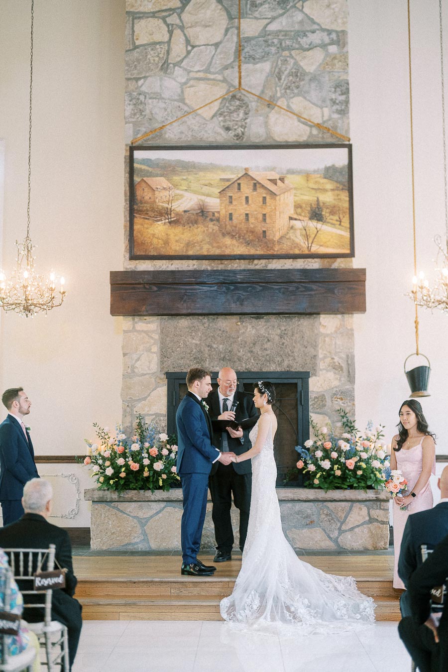 Miffy and Rawle hold hands during their chapel ceremony before a stone fireplace with florals and attendants.