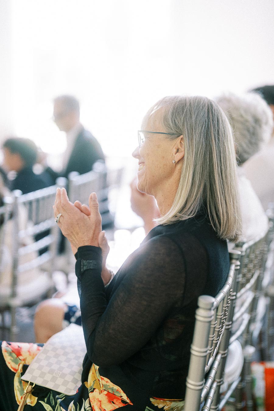 Smiling woman with glasses applauds while seated on a silver chair among wedding guests.