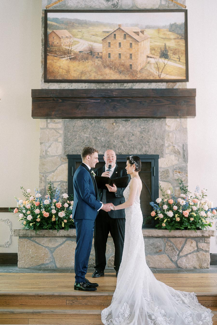 Miffy and Rawle hold hands during their indoor wedding ceremony before a stone fireplace and floral arrangements.