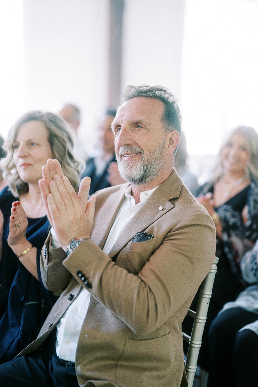 Smiling man in a tan blazer claps while seated among wedding guests during an indoor celebration.
