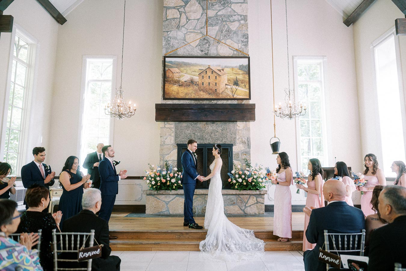Bride and groom hold hands during their chapel ceremony as wedding party and guests applaud beside a stone fireplace.