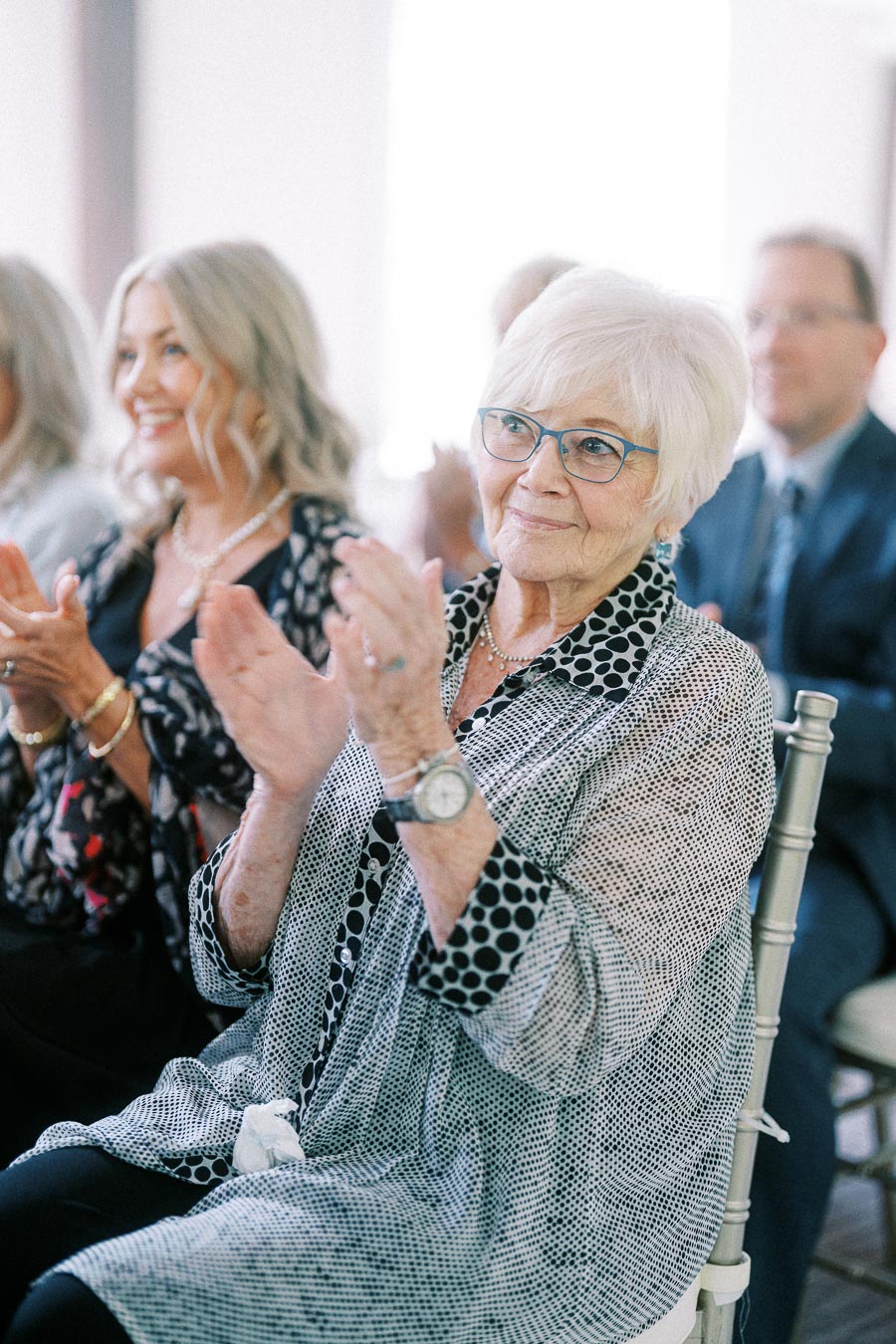 Elderly guest in blue glasses clapping while seated among wedding guests during an indoor ceremony