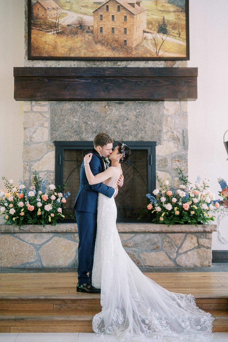 Miffy and Rawle kiss in wedding attire before a stone fireplace with floral arrangements.