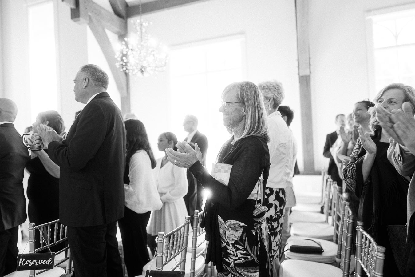 Wedding guests stand and applaud beside silver chairs in a bright ceremony room at Ancaster Mill.