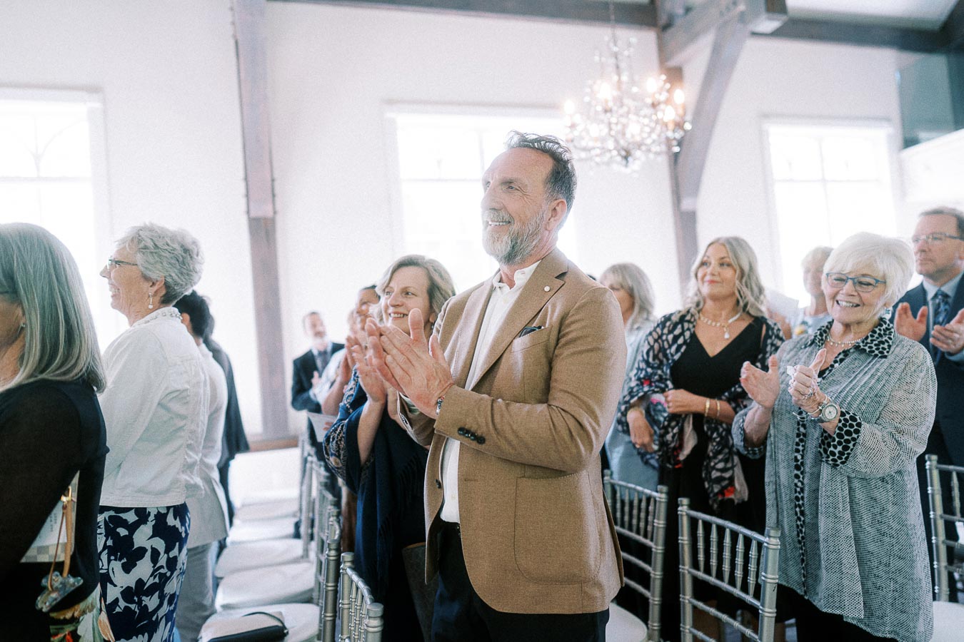 Wedding guests stand and clap among white chairs in a bright ceremony room with chandeliers.