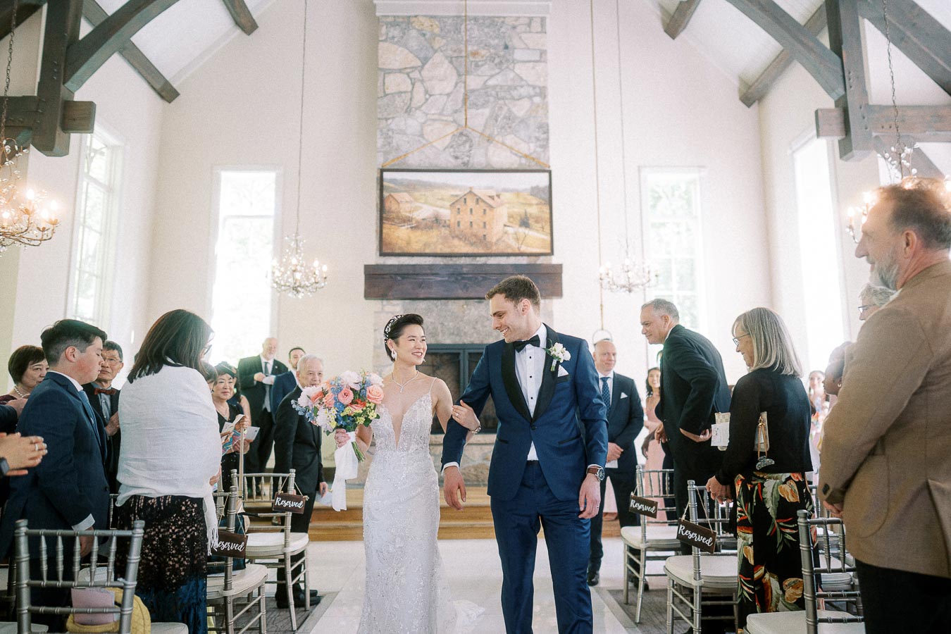 Miffy and Rawle walk down the chapel aisle smiling after their wedding ceremony as guests stand beside them.