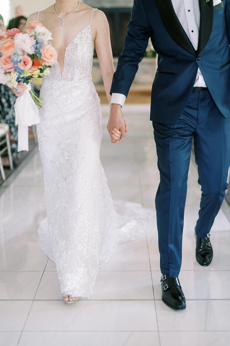 Bride and groom walk hand in hand, showing a lace wedding dress, navy tuxedo, and pastel bouquet.