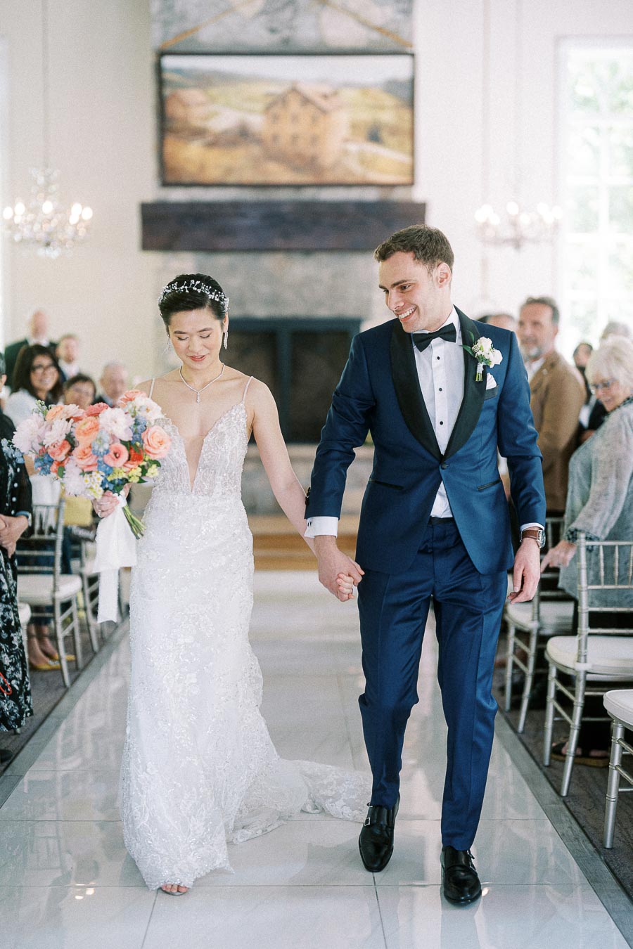 Miffy and Rawle walk hand in hand down the ceremony aisle, holding a pastel bouquet as seated guests watch.