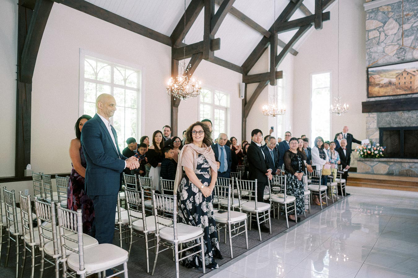 Wedding guests stand beside silver chairs in a bright chapel with timber beams and chandeliers.