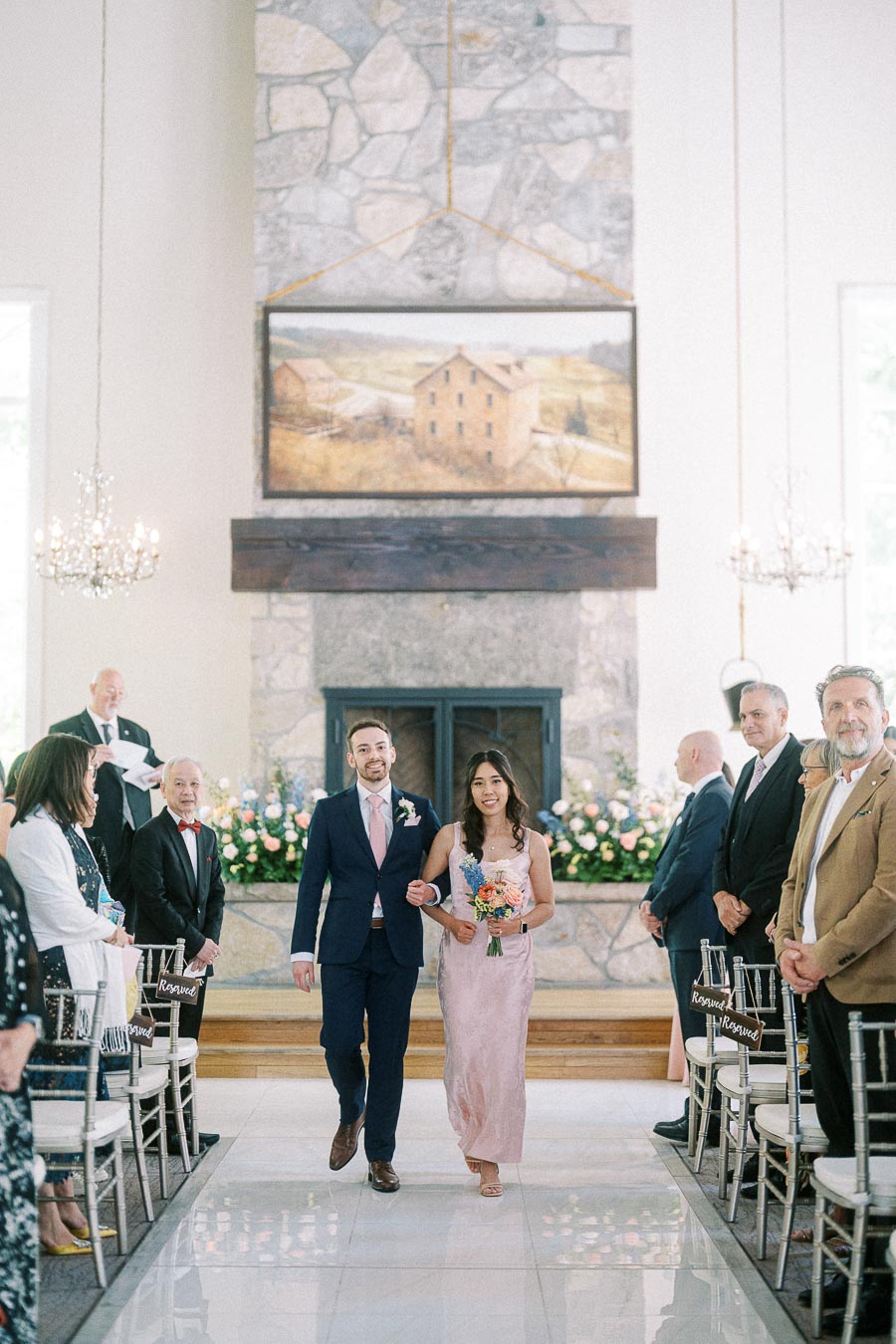 Bridesmaid and groomsman walk arm in arm down the aisle in a bright stone fireplace ceremony room.
