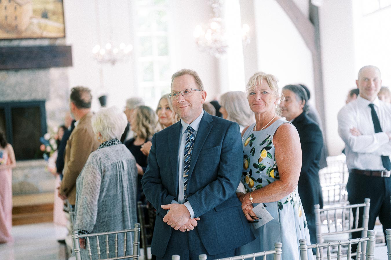 Wedding guests stand between chairs in a bright ceremony room with chandeliers and a stone fireplace.