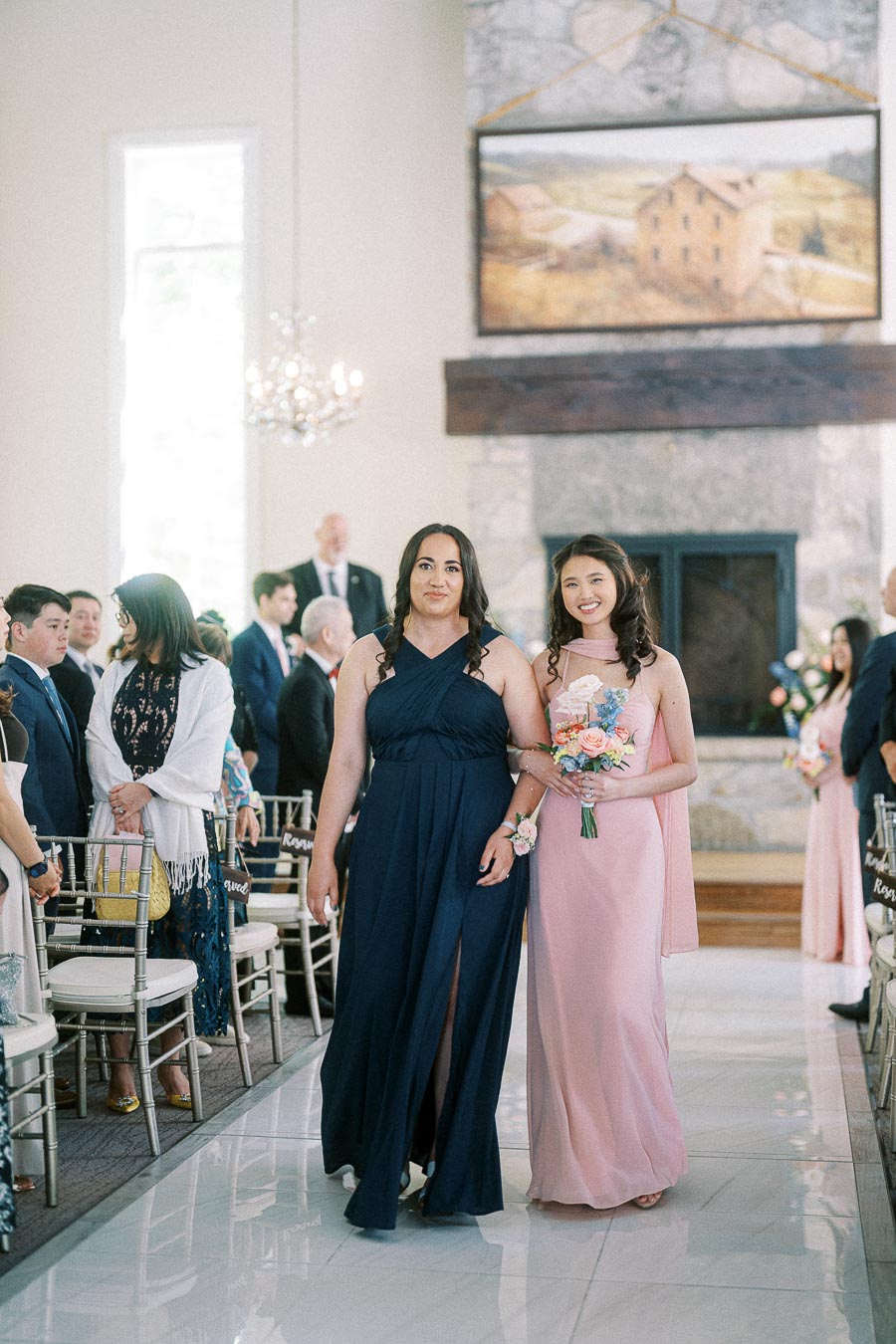 Two bridesmaids in navy and blush dresses walk down the chapel aisle with a small bouquet as guests stand nearby.
