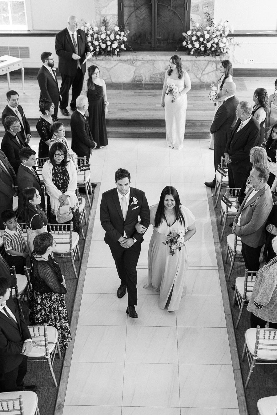 Wedding party pair walks down the ceremony aisle between seated guests, with bride and attendants waiting at the front.