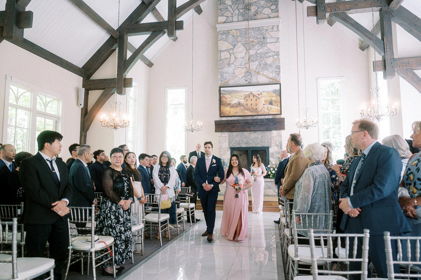 Wedding party pair walks down the aisle as guests stand in a timber-framed chapel at Ancaster Mill.