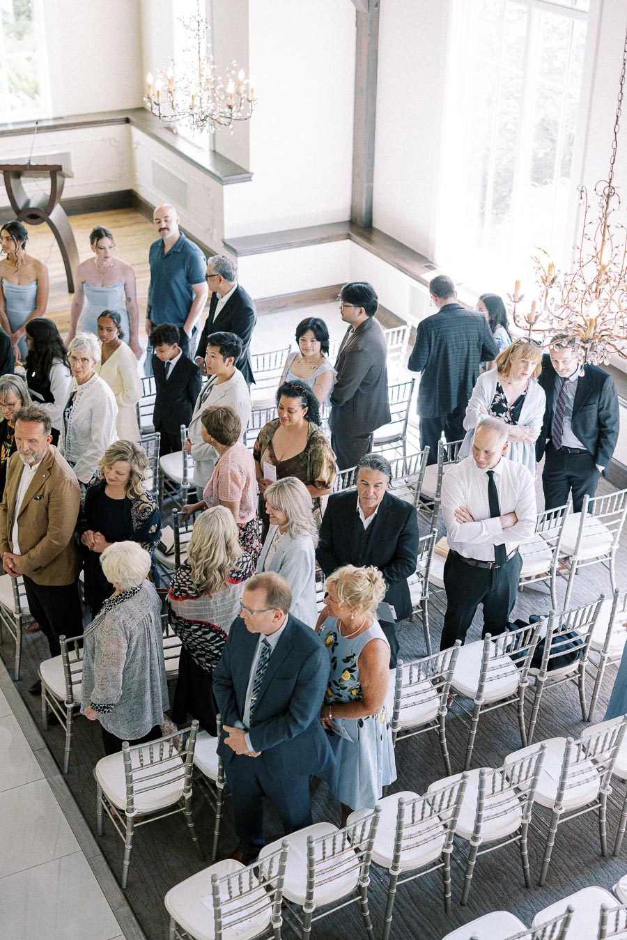 Wedding guests stand among silver chairs in a bright chapel with chandeliers before the ceremony.