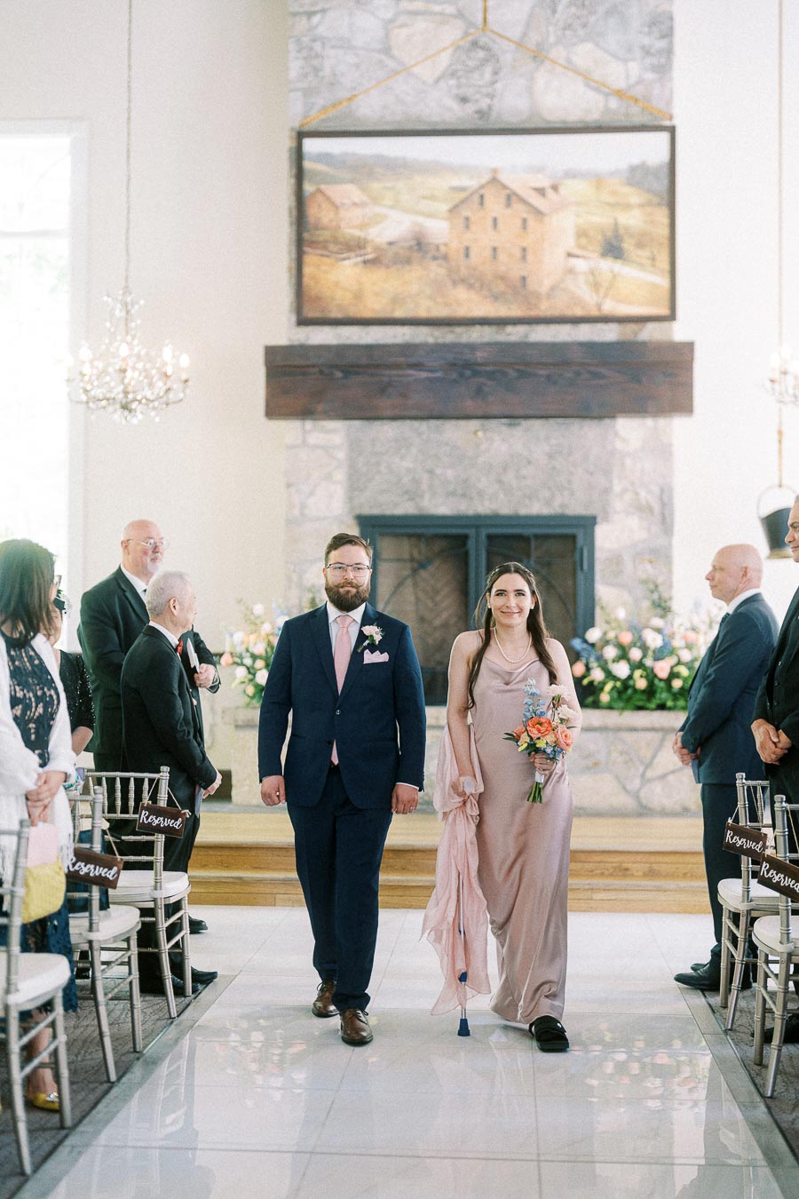 Groomsman and bridesmaid walk down the chapel aisle between seated guests at Ancaster Mill.