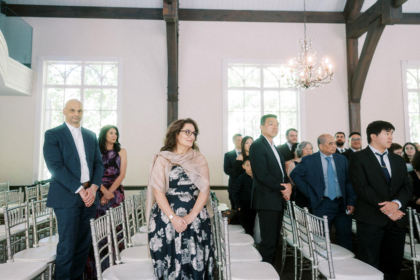 Wedding guests stand between white ceremony chairs in a bright chapel with chandeliers and wooden beams.