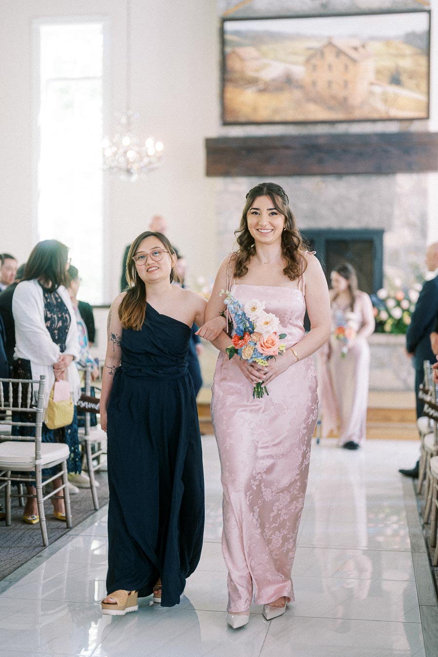Two women walk arm in arm down the wedding aisle, one holding a colorful bouquet in a bright chapel space.