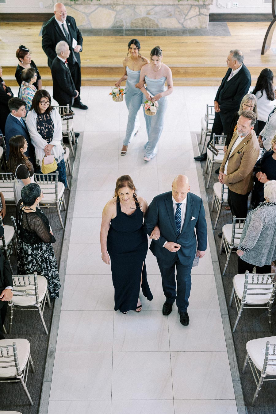 Wedding processional with paired attendants walking down the aisle as guests stand beside silver chairs.