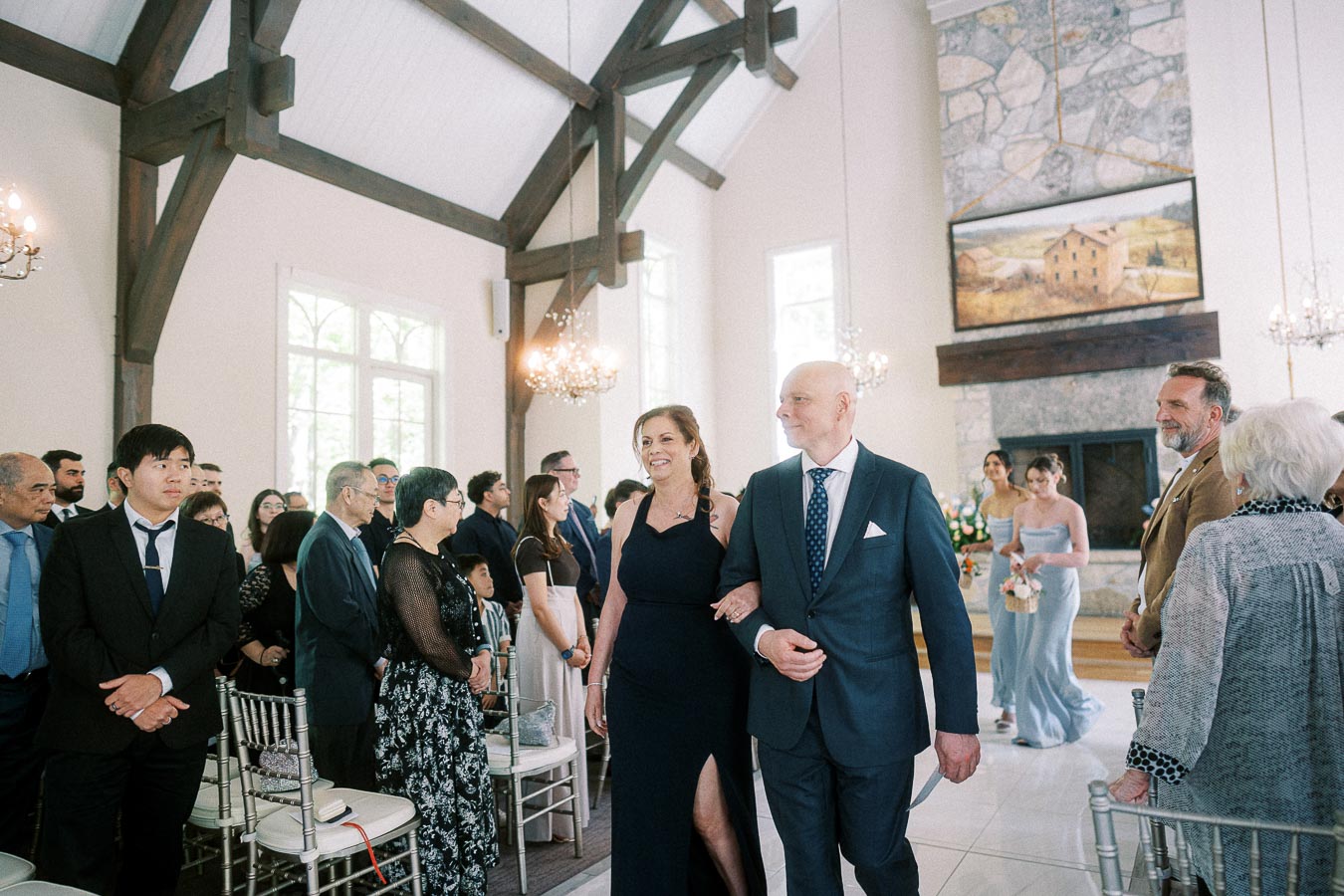 A formally dressed couple walks arm in arm down the chapel aisle as wedding guests stand beside white chairs.