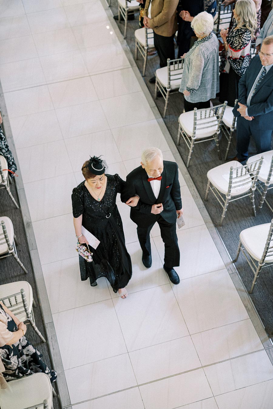 Overhead view of an older couple walking arm in arm down a white wedding aisle as guests stand beside silver chairs.