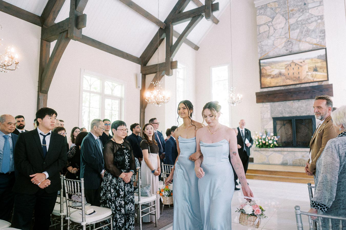 Two bridesmaids in pale blue dresses walk down the chapel aisle carrying flower baskets as guests stand.
