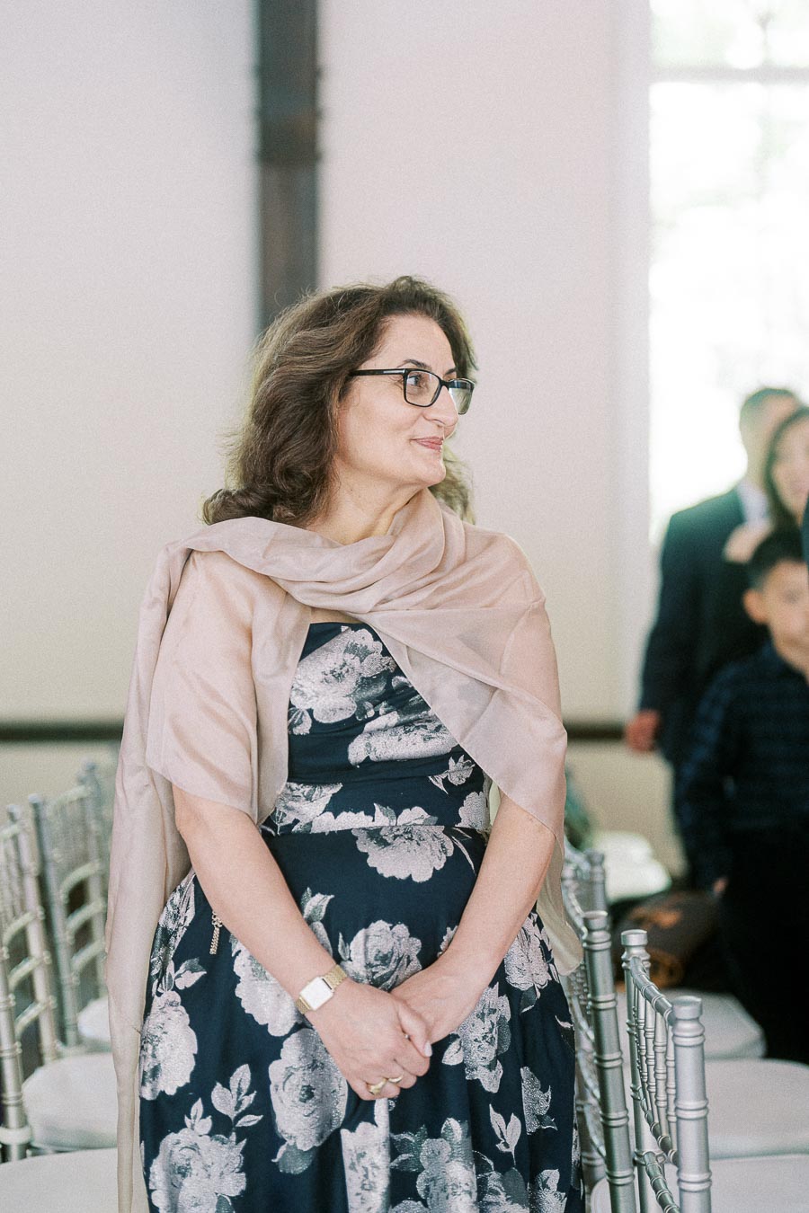 Woman in glasses and a floral dress stands between ceremony chairs, looking to the side in a bright indoor venue.