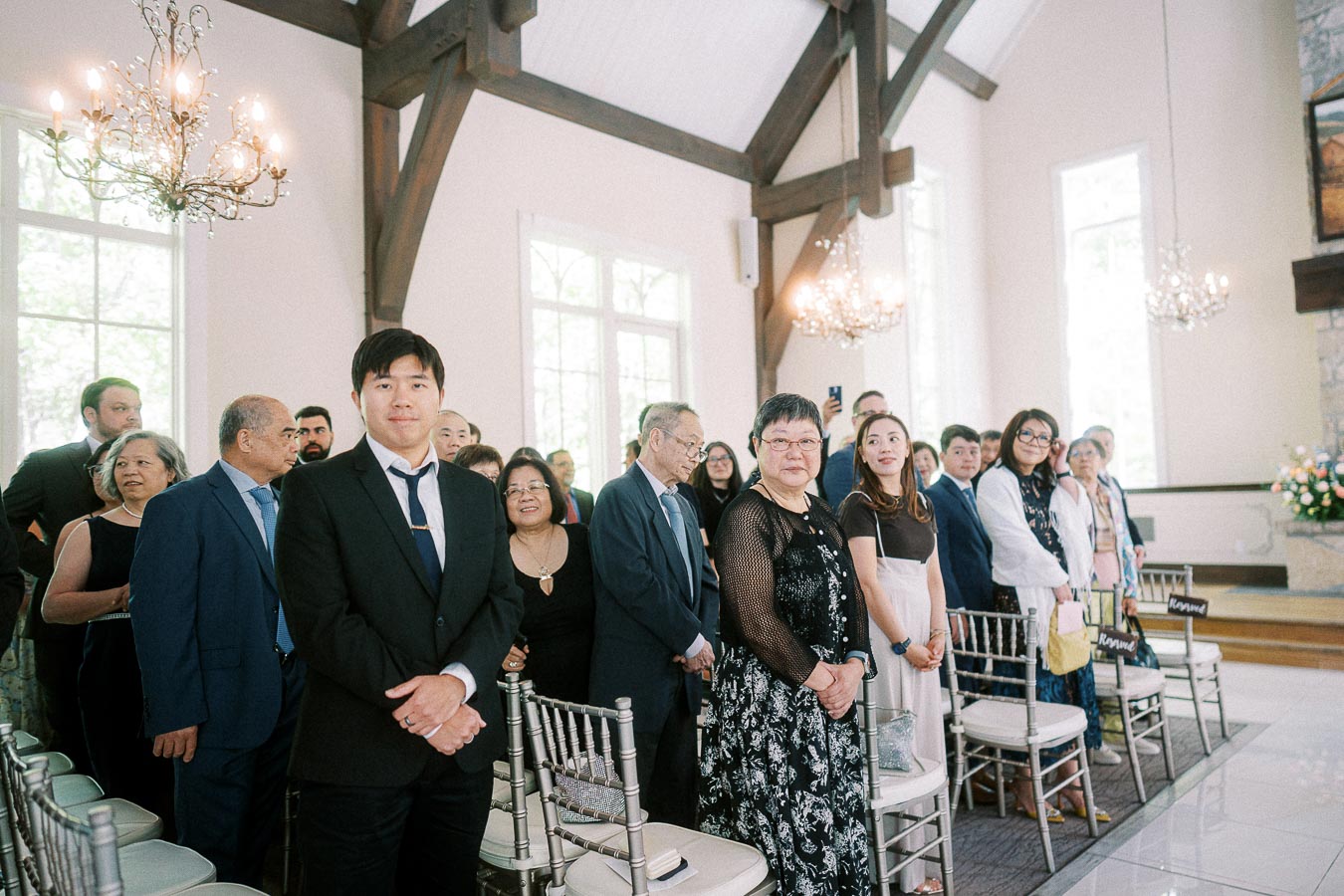 Wedding guests stand beside silver chairs in a bright chapel with chandeliers and wooden beams.