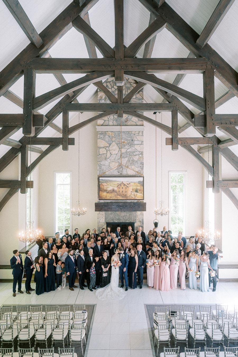 Large wedding group portrait in a chapel with timber beams, stone fireplace, chandeliers, and rows of chairs.