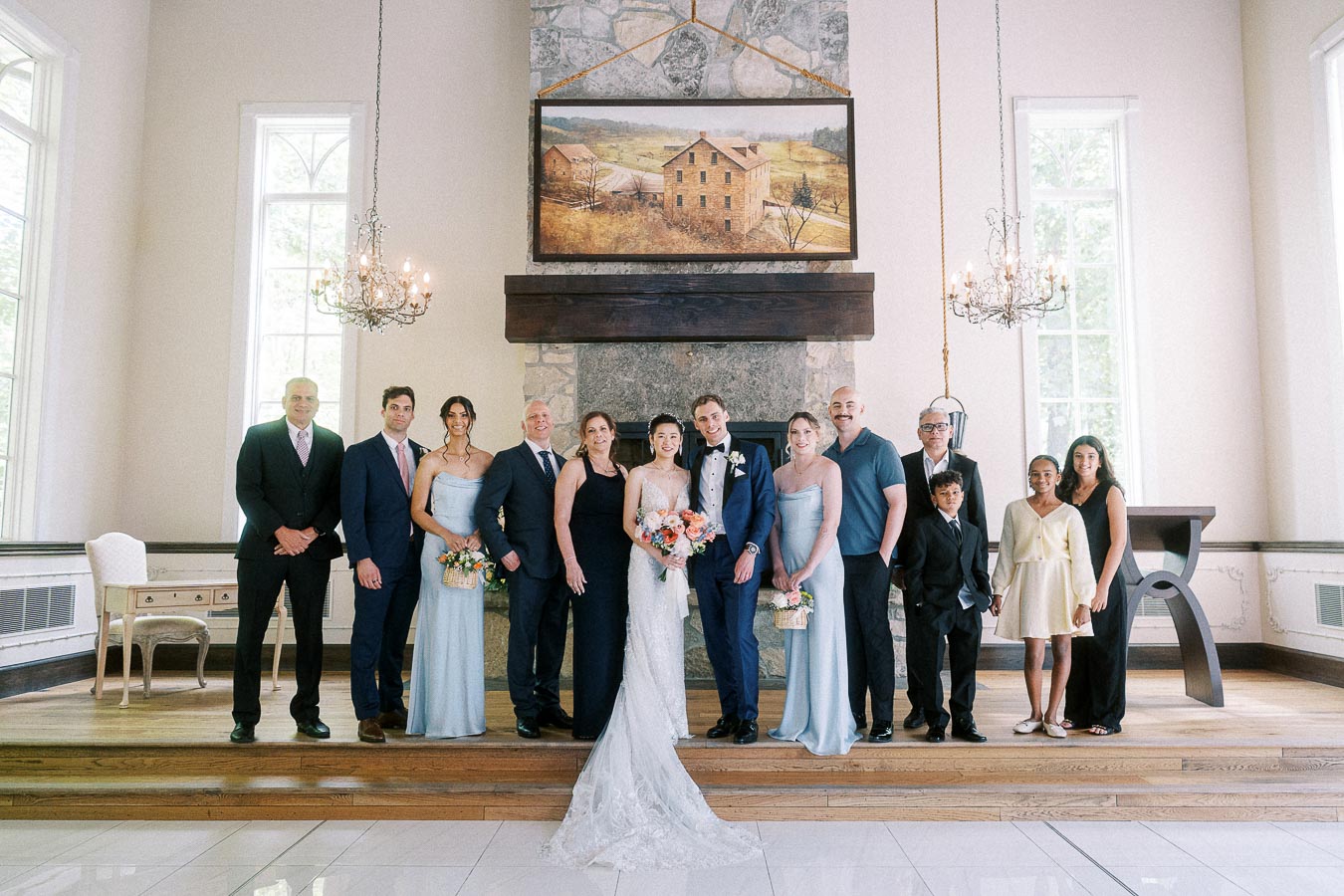 Miffy and Rawle stand with family and attendants in formal wear before a stone fireplace and chandeliers.