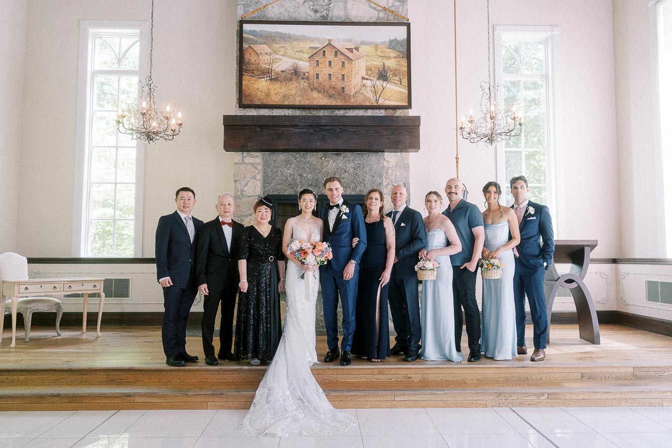 Miffy and Rawle pose with family and wedding party in a bright Ancaster Mill room with chandeliers and a fireplace.