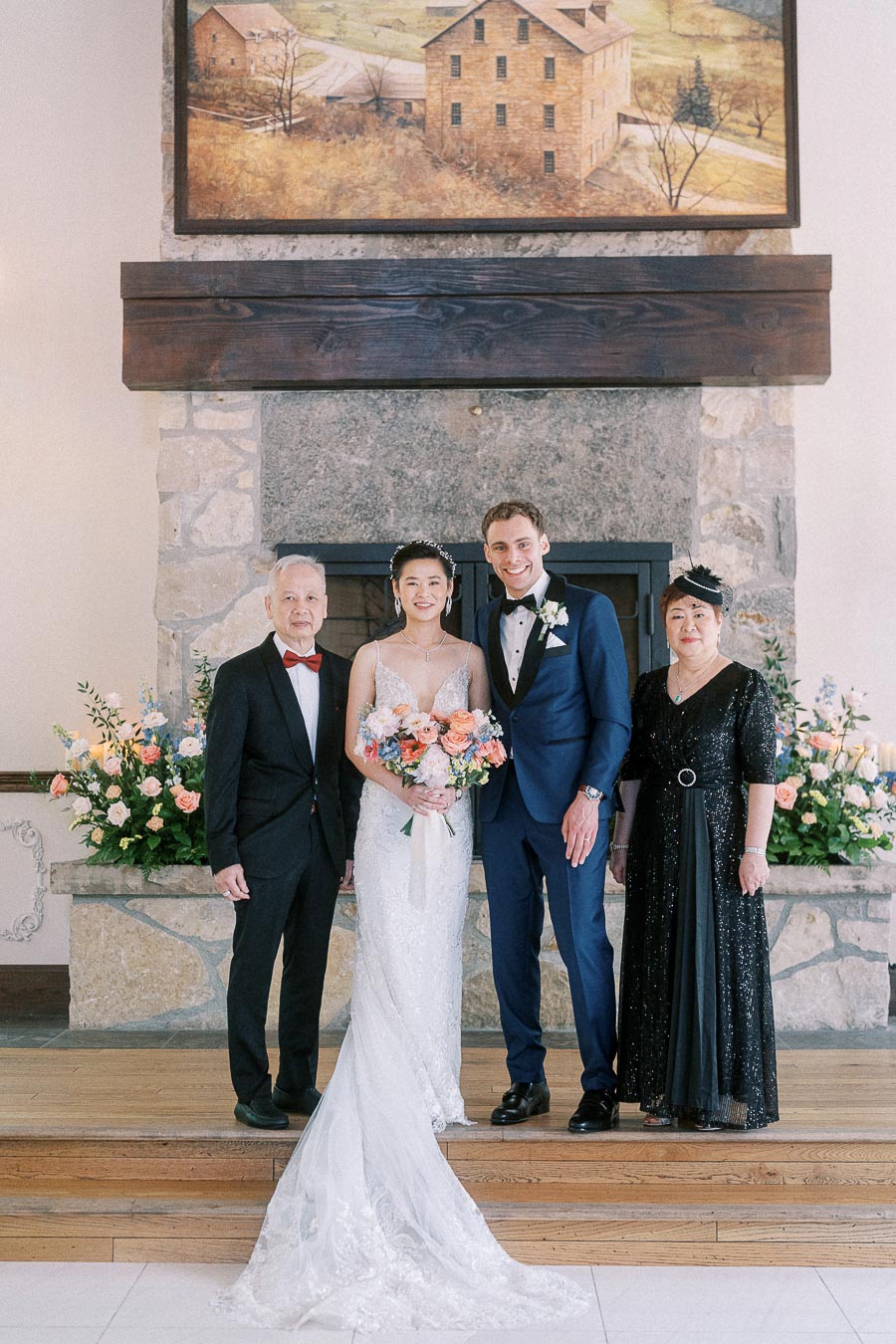 Miffy and Rawle stand with two family members in formal attire before a stone fireplace and floral arrangements.