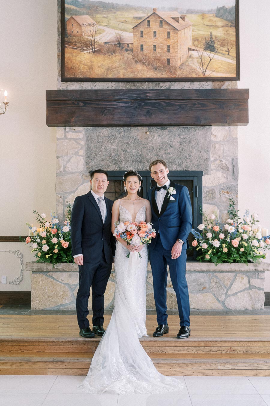 Miffy stands in her wedding dress between Rawle and a suited man in front of a stone fireplace with flowers.