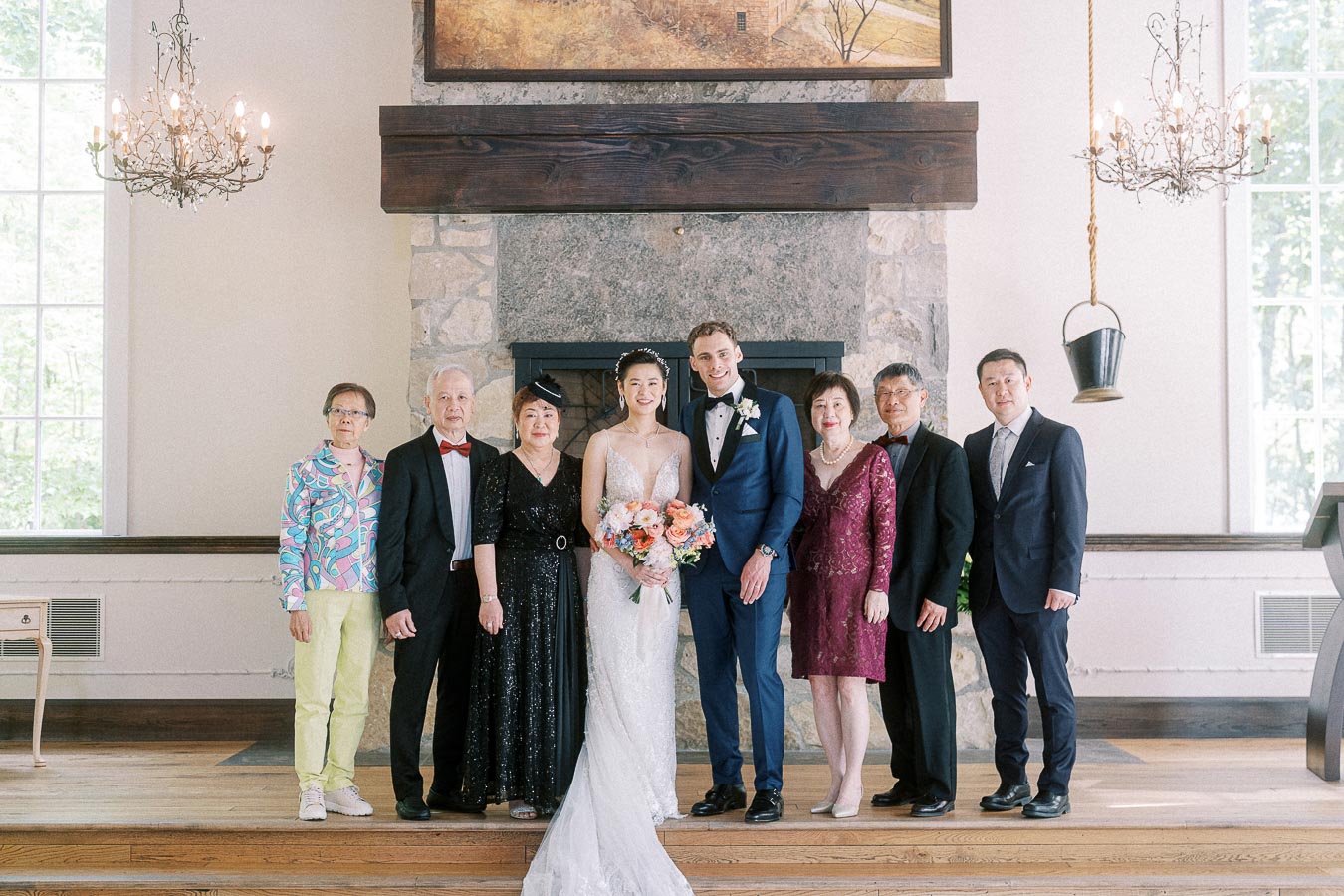 Bride and groom stand with six relatives for a formal family portrait in front of a stone fireplace.