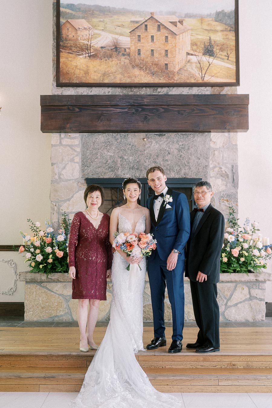 Miffy and Rawle stand with two parents before a stone fireplace with floral arrangements at their wedding.