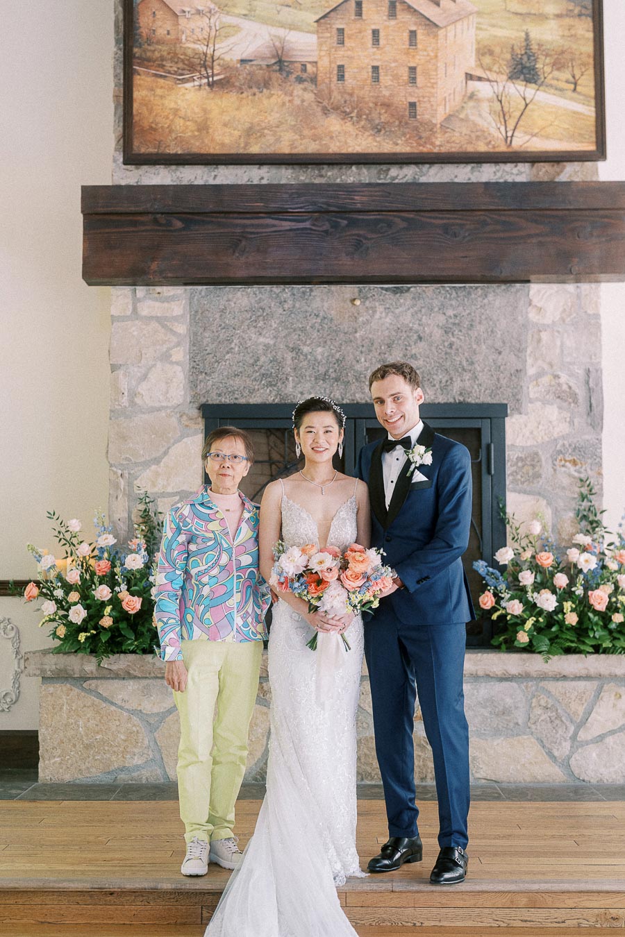 Bride and groom pose with an older woman in front of a stone fireplace decorated with pastel wedding flowers.
