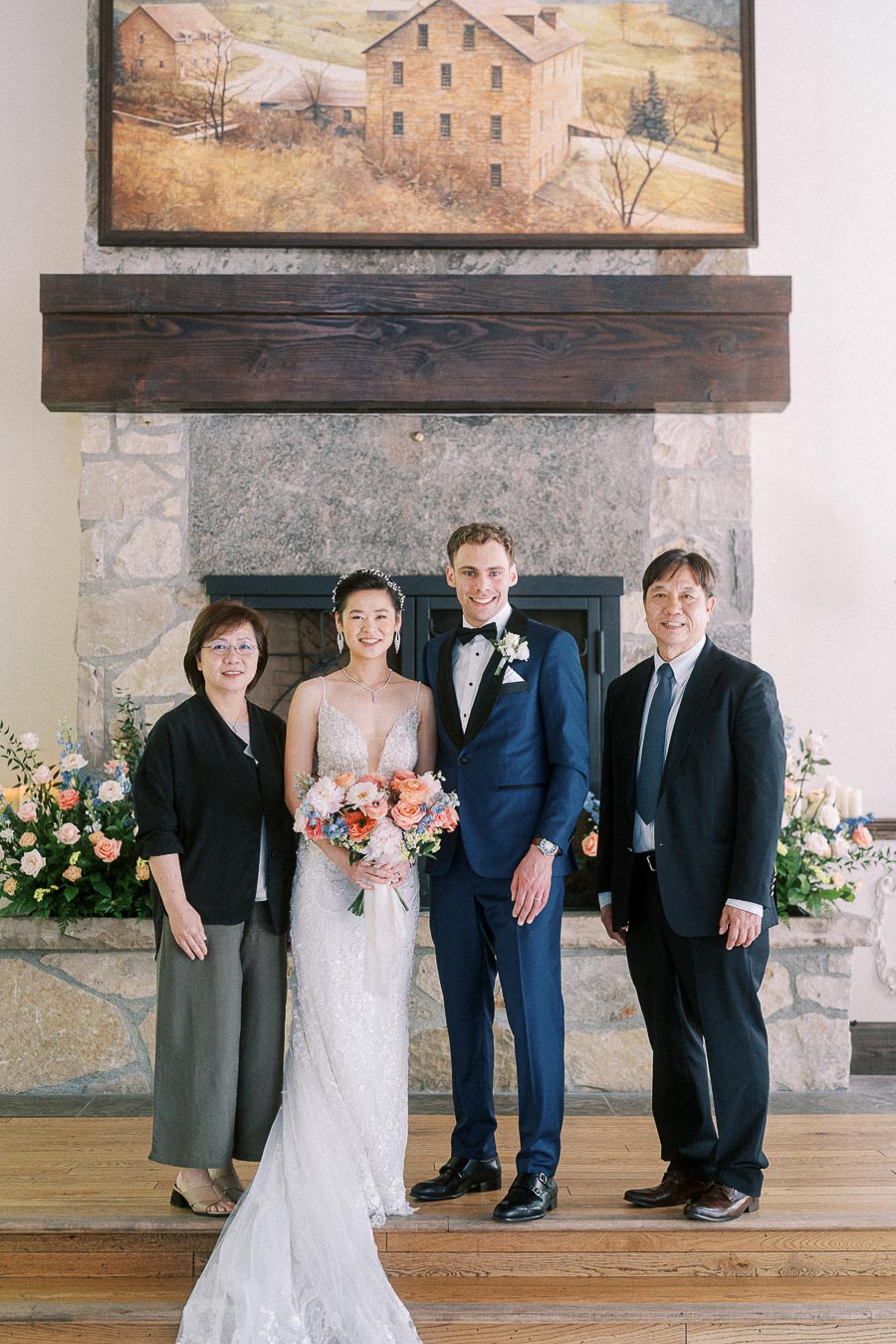 Bride and groom stand with two parents in front of a stone fireplace for a formal wedding portrait.