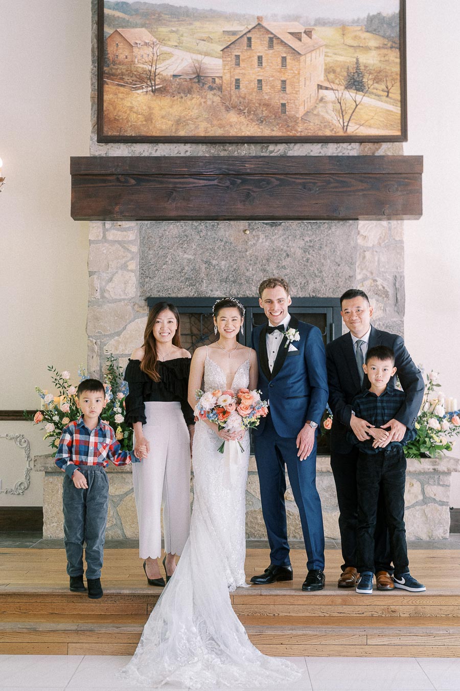 Miffy and Rawle pose with family by a stone fireplace at Ancaster Mill, with floral arrangements and children beside them.