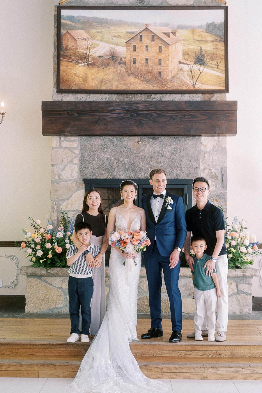 Bride and groom pose with two adults and two children in front of a stone fireplace at Ancaster Mill.