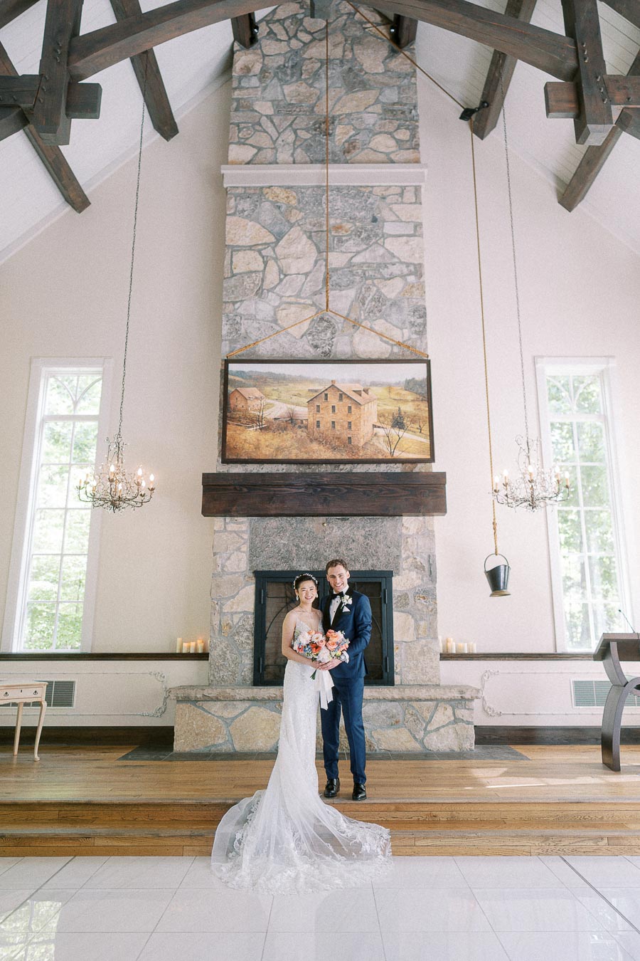 Bride and groom stand before a stone fireplace inside a chapel-like room with chandeliers and wood beams.