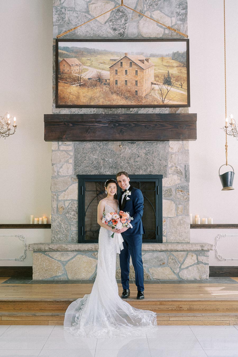 Miffy and Rawle stand together in wedding attire before a stone fireplace at Ancaster Mill.